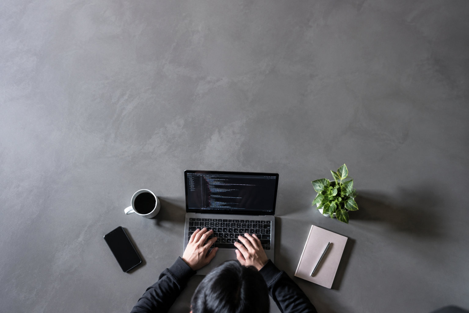 Vue de dessus d'une personne codant sur un ordinateur portable, entourée d'un smartphone, d'un café et d'une plante.